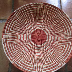 May include: A round, shallow woven basket featuring a coiled construction with a geometric pattern in alternating shades of terracotta orange and cream. The design radiates from a solid circular center, expanding outward in a symmetrical maze-like motif.