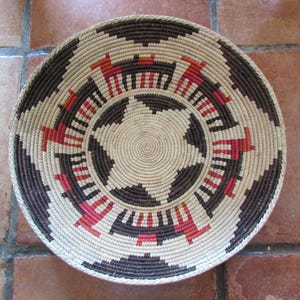 May include: A round woven basket featuring a coiled design with a central star pattern. The basket displays a repeating motif of stylized animals in alternating dark brown and red colors against a natural light beige fiber background.