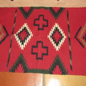 May include: Red, black, white, and green woven rug with a geometric pattern. The rug has a fringe on the ends.