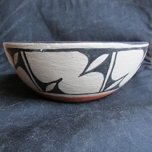 May include: A round ceramic bowl featuring a matte cream-colored exterior decorated with sharp, black geometric patterns. The rim is painted with a thin black line, while the base of the bowl reveals a contrasting terracotta red clay finish.