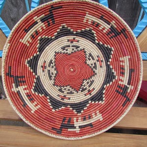 May include: A round, coiled basket tray featuring a geometric pattern in red, black, and cream. The center displays a star-like motif surrounded by concentric circles, while the outer border is decorated with stylized animal figures in black and cream.