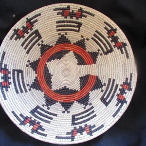 May include: A woven basket with a circular design in shades of white, black, and red. The basket has a central circle with concentric rings of black and red.