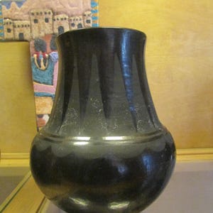 May include: A black ceramic vase with a rounded body and a flared rim. The surface features a matte black finish with a series of vertical, triangular etched patterns around the neck and a scalloped band design encircling the widest part.