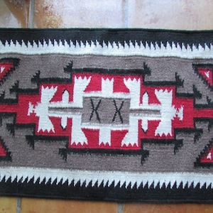 May include: A Native American rug with a geometric design in red, white, and black on a gray background. The rug has a black border with a white fringe.