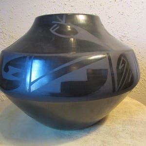 May include: A black ceramic vase with a rounded base and a flared rim. The vase features geometric designs in a lighter gray color, creating a contrast against the dark background. The vase is sitting on a light-colored surface.