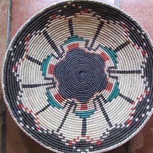May include: A round woven basket with a central spiral design in dark blue. The basket features a repeating pattern of black, red, and teal accents against a cream-colored background. The rim is dark blue.