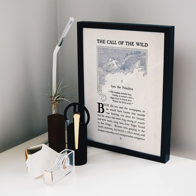 May include: A framed print of the first page of "The Call of the Wild" by Jack London. The text is in black and white and features a map of a mountainous region. The frame is black and the print is on white paper.