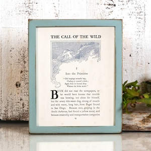 May include: A framed print of the first page of "The Call of the Wild" by Jack London. The print features a blue and white illustration of a mountain range and the text "Into the Primitive". The frame is a light blue color.