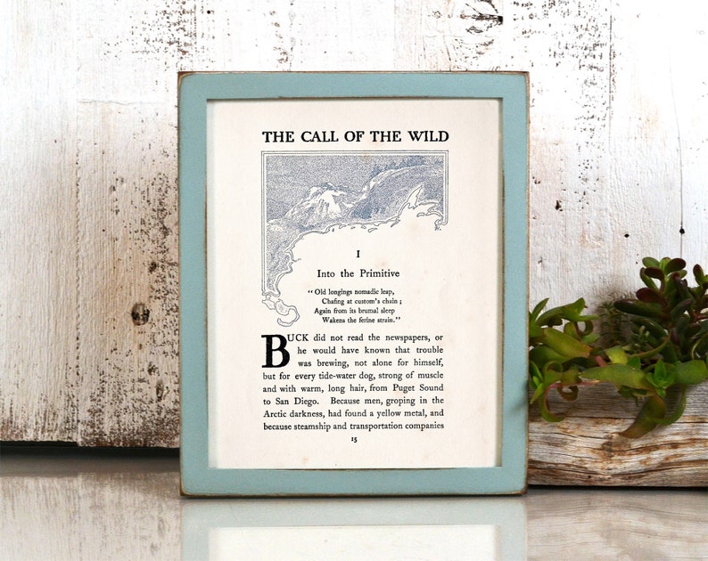 May include: A framed print of the first page of "The Call of the Wild" by Jack London. The print features a blue and white illustration of a mountain range and the text "Into the Primitive". The frame is a light blue color.