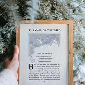 May include: A framed print of a book excerpt from "The Call of the Wild" by Jack London. The excerpt is from the first chapter, "Into the Primitive". The text is printed in black ink on a white background. The frame is made of wood and has a light brown finish.