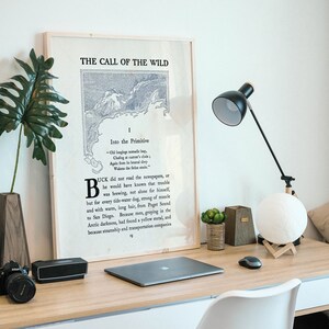 May include: A framed print of the first page of "The Call of the Wild" by Jack London. The print is on a wooden desk with a lamp, a moon lamp, a laptop, a mouse, and a potted plant.