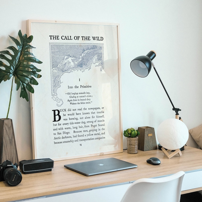 May include: A framed print of the first page of "The Call of the Wild" by Jack London. The print is on a wooden desk with a lamp, a moon lamp, a laptop, a mouse, and a potted plant.