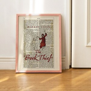 Pode incluir: Uma impress&atilde;o emoldurada de uma capa de livro com o t&iacute;tulo "The Book Thief" em letras vermelhas e pretas. O fundo &eacute; uma p&aacute;gina de dicion&aacute;rio vintage com a palavra "Death" no topo.