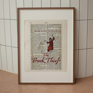 Pode incluir: Uma impress&atilde;o emoldurada de uma capa de livro com uma ilustra&ccedil;&atilde;o vermelha e preta de uma figura com uma t&uacute;nica com capuz segurando uma foice. O t&iacute;tulo do livro &eacute; "The Book Thief" em letras vermelhas.