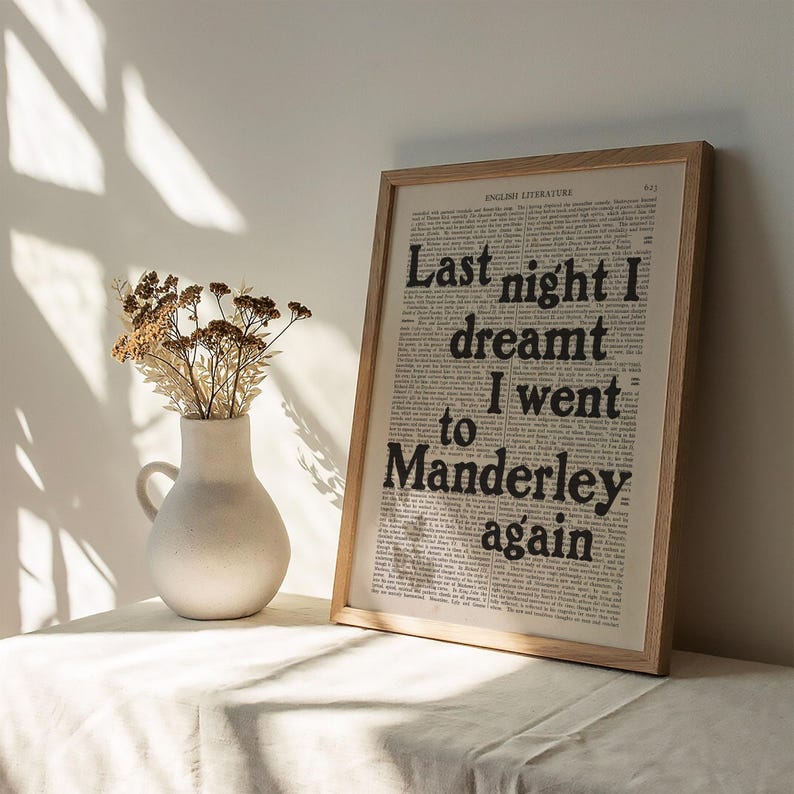 May include: A framed print with a quote from the novel "Rebecca" by Daphne du Maurier. The quote reads "Last night I dreamt I went to Manderley again". The print is on a white surface with a vase of dried flowers.