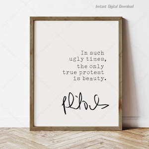 May include: A printable quote in a black frame on a wooden floor. The quote reads "In such ugly times, the only true protest is beauty." with the word "beauty" underlined. The signature "flos" is written in cursive below the quote.