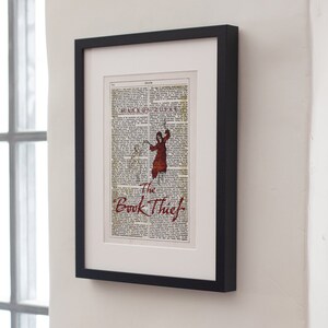 Pode incluir: Uma impress&atilde;o emoldurada de uma p&aacute;gina de livro com uma ilustra&ccedil;&atilde;o vermelha de uma pessoa com um capuz. O texto "The Book Thief" est&aacute; impresso em vermelho na parte inferior da p&aacute;gina.