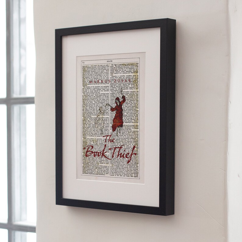 Pode incluir: Uma impress&atilde;o emoldurada de uma p&aacute;gina de livro com uma ilustra&ccedil;&atilde;o vermelha de uma pessoa com um capuz. O texto "The Book Thief" est&aacute; impresso em vermelho na parte inferior da p&aacute;gina.