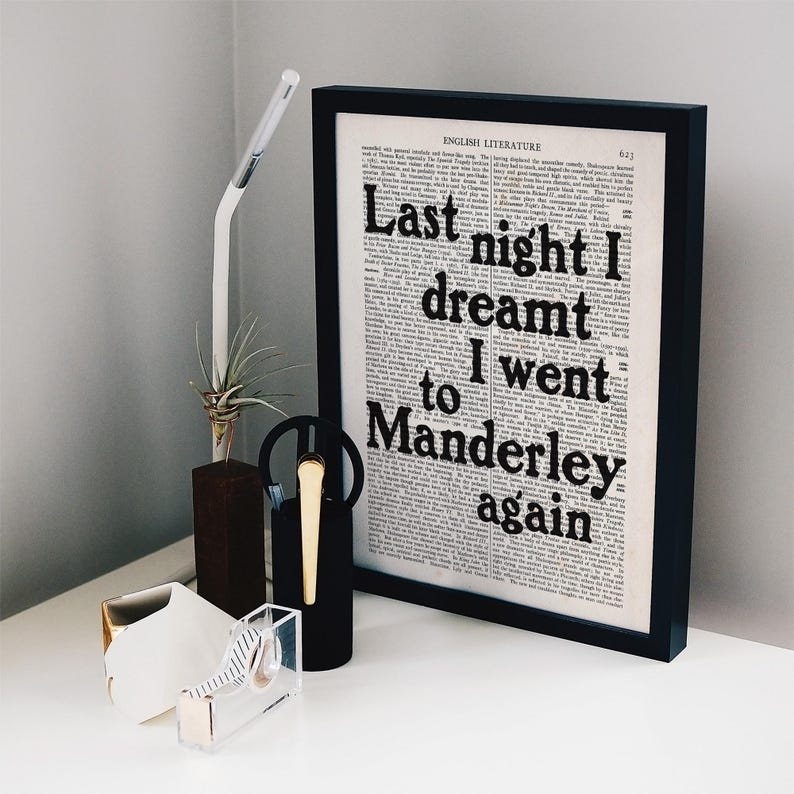 May include: A black framed print with the text "Last night I dreamt I went to Manderley again" on a white background. The print is sitting on a white surface with a small plant, a gold and clear tape dispenser, and a black and gold pen holder.