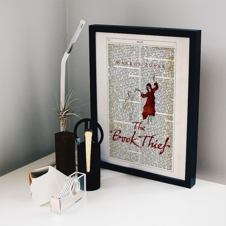 Pode incluir: Uma impress&atilde;o emoldurada preta com um fundo de p&aacute;gina de livro vintage apresentando uma figura vermelha em um manto com capuz. O texto "The Book Thief" est&aacute; escrito em letras cursivas vermelhas.