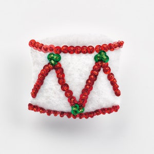 Puede incluir: Un tambor de fieltro blanco decorado con lentejuelas rojas y verdes. El tambor tiene un diseño simple de dos triángulos.
