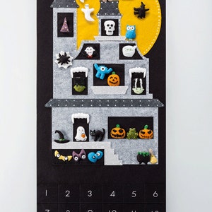 Pode incluir: Um calend&aacute;rio do advento de feltro em forma de casa assombrada com uma lua amarela e v&aacute;rias decora&ccedil;&otilde;es de feltro. O calend&aacute;rio tem os n&uacute;meros de 1 a 30.