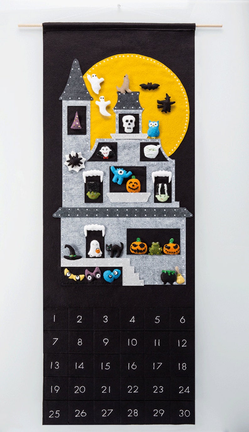 Pode incluir: Um calend&aacute;rio do advento de feltro em forma de casa assombrada com uma lua amarela e v&aacute;rias decora&ccedil;&otilde;es de feltro. O calend&aacute;rio tem os n&uacute;meros de 1 a 30.
