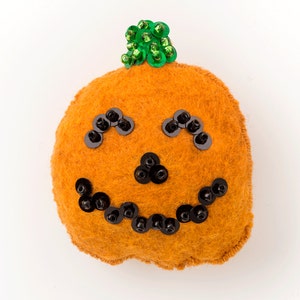 Pode incluir: Uma abóbora laranja de feltro com um caule de lantejoulas verdes e um rosto de lantejoulas pretas. A abóbora tem uma expressão sorridente com olhos, nariz e boca.