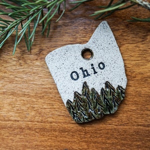 May include: White ceramic ornament shaped like the state of Ohio with a green and brown tree design and the word "Ohio" written in black.