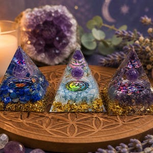May include: Three pyramid-shaped orgone energy generators with embedded crystals and gold flakes. Each pyramid features a different color scheme: blue, light blue, and purple. The pyramids sit on a wooden tray with a floral design, with a lit candle and amethyst geode in the background.