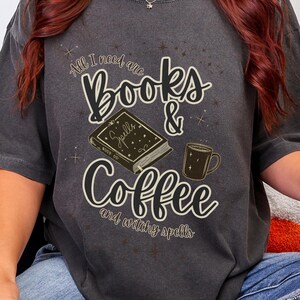 Op de afbeelding: Een antracietgrijs T-shirt met korte mouwen en een grafische print met de tekst All I need are Books & Coffee and witchy spells, een illustratie van een boek met de titel Spells en een koffiemok. Gecombineerd met een blauwe spijkerbroek.