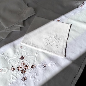 Op de afbeelding: Witte linnen tafelkleed en servetten met geborduurde bloemen en geometrische ontwerpen. Het tafelkleed heeft een groot centraal patroon met uitgesneden details en een decoratieve rand. Servetten zijn links opgestapeld.