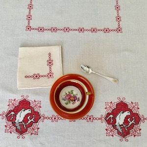 Pode incluir: Um conjunto de chá vintage com um pires vermelho e uma chávena floral. Uma pequena colher de prata e um guardanapo de linho são colocados sobre uma toalha de mesa branca com detalhes bordados a vermelho. A toalha de mesa apresenta um padrão repetitivo e uma borda decorativa.