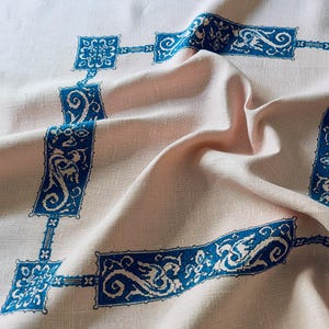 Pode incluir: Uma toalha de mesa de linho rosa claro com um padrão repetitivo de bordas azuis ornamentadas e detalhes quadrados. O tecido é suavemente drapeado, mostrando a textura e o design do tecido.