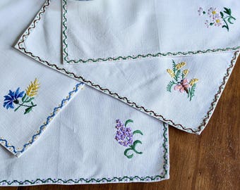 Op de afbeelding: Witte linnen placemats met geborduurde bloemmotieven. Elke placemat heeft een andere bloem, waaronder blauwe, gele, roze en paarse bloemen. De randen zijn afgewerkt met een decoratieve gestikte rand. Een kop en schotel zijn zichtbaar op de achtergrond.