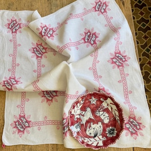 以下が含まれることがあります： ピンク色の刺繍が施された鳥のモチーフが線状のデザインで繋がった白いリネンクロス。縁が波打った円形の陶器製プレートがクロスの上に置かれており、黒い線画で描かれた象のイラストが描かれています。