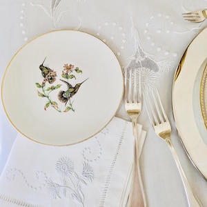 May include: A white plate with a gold rim features a detailed illustration of two hummingbirds and flowers. Silver forks and a white embroidered napkin are arranged on a white tablecloth, alongside a second plate.
