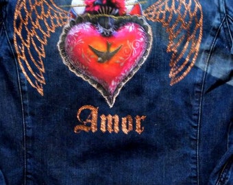 May include: A blue denim jacket with a painted design of a heart with wings and the word "Amor" in gold glitter.