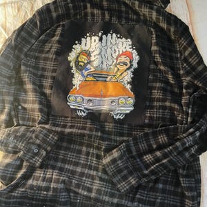 May include: Black and gray plaid flannel shirt with a graphic on the back. The graphic features two cartoon figures in a vintage orange car with the text "UP IN SMOKE". The shirt has buttoned cuffs and a collar.