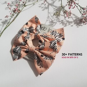 May include: A peach-coloured hair bow with a zebra print pattern. The bow is made of fabric and has a simple design. The text "30+ PATTERNS SOLD IN SETS OF 2" is printed on a white background.