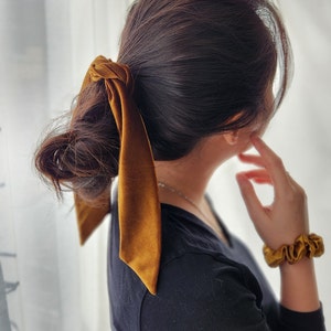 May include: A woman with long brown hair tied back with a mustard yellow satin scrunchie. She is wearing a black long-sleeved shirt and a mustard yellow scrunchie on her wrist.
