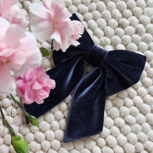 May include: A navy blue velvet bow with a large, centered knot. The bow is lying on a white, textured surface.