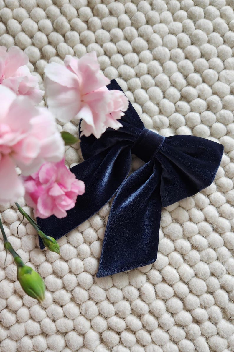 May include: A navy blue velvet bow with a large, centered knot. The bow is lying on a white, textured surface.
