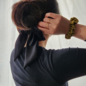 May include: A person with dark brown hair wearing a black long-sleeved shirt with a black satin ribbon tied in a bow at the back of the neck. The person is wearing a gold ring on their finger and a green scrunchie on their wrist.