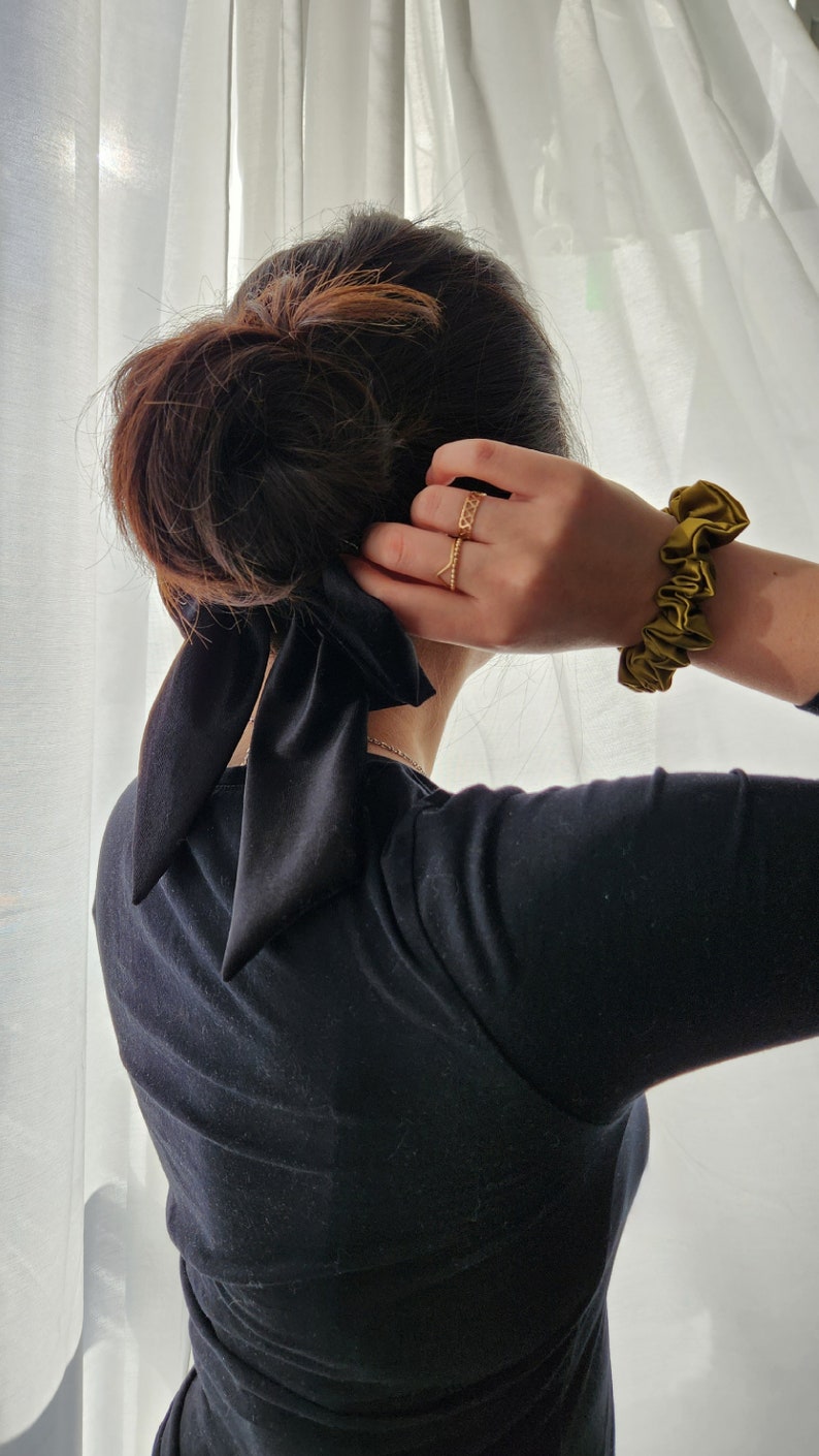 May include: A person with dark brown hair wearing a black long-sleeved shirt with a black satin ribbon tied in a bow at the back of the neck. The person is wearing a gold ring on their finger and a green scrunchie on their wrist.