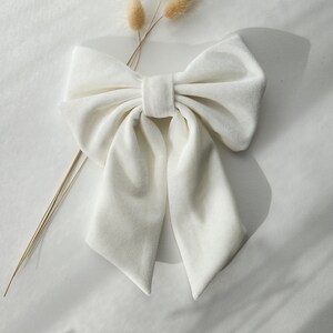 May include: A large white velvet bow with a center loop. The bow is on a white surface with a dried flower sprig.