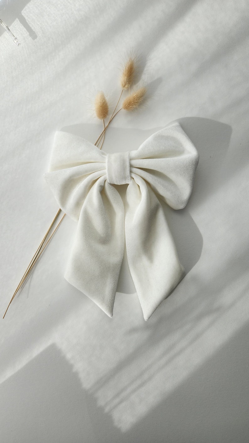 May include: A large white velvet bow with a center loop. The bow is on a white surface with a dried flower sprig.