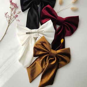 May include: Four velvet hair bows in different colors: black, white, burgundy, and brown. The bows are tied with a simple knot and have a clip on the back.