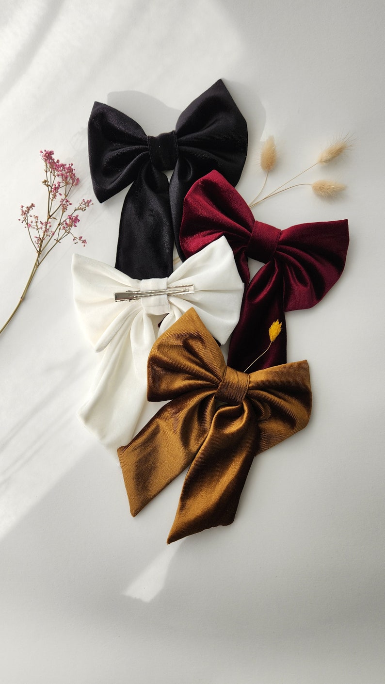 May include: Four velvet hair bows in different colors: black, white, burgundy, and brown. The bows are tied with a simple knot and have a clip on the back.