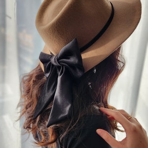 May include: A brown felt fedora hat with a black velvet ribbon and a large black satin bow. The hat is worn by a person with long brown hair.